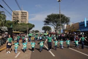 Crianças e adultos de mãos dadas participam de desfile em Dourados, com público nas laterais da avenida. Foto de arquivo.